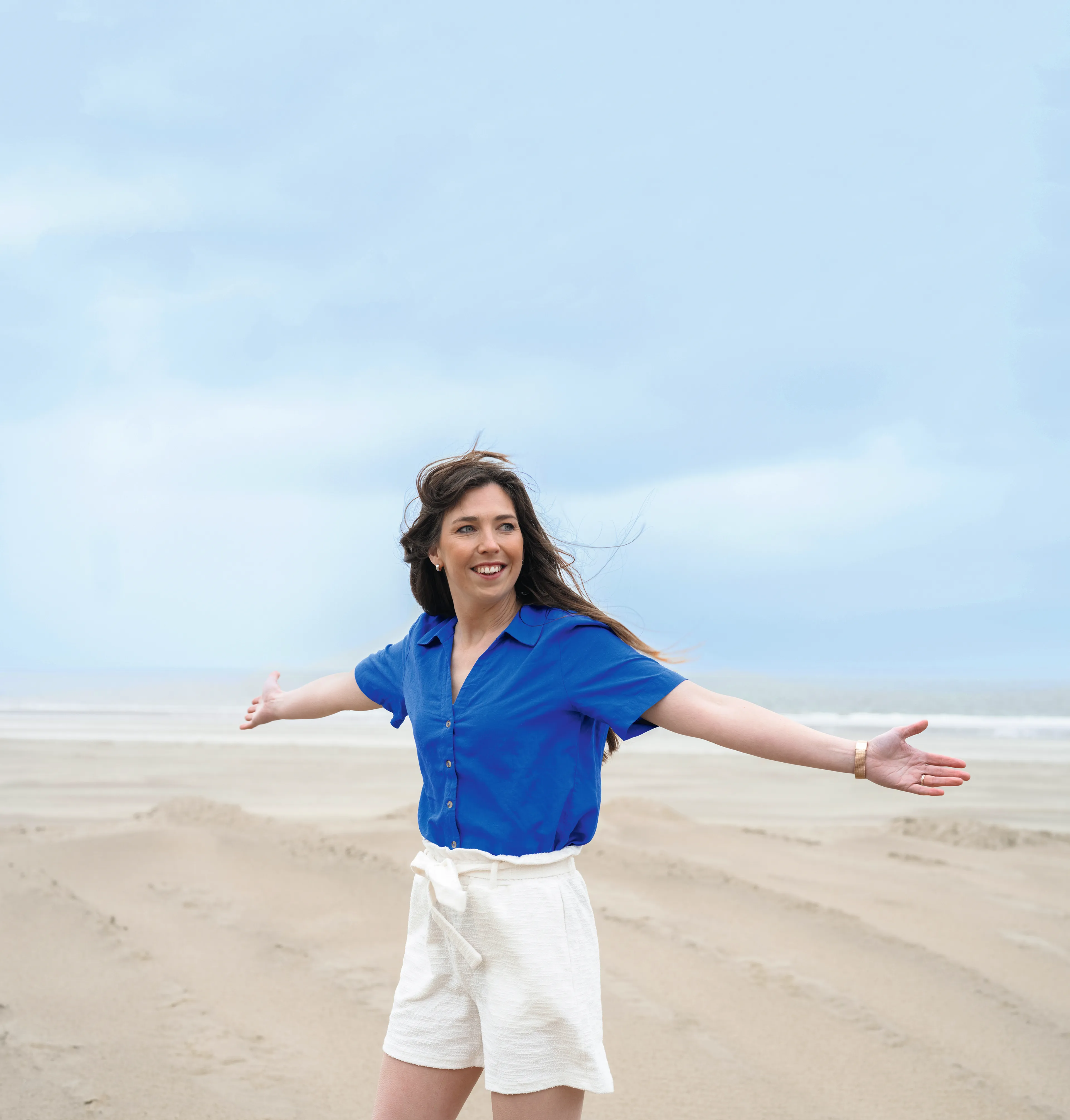 Op een breed zandstrand staat een jongvolwassen vrouw met een glimlach en open armen, terwijl haar lange, donkere haar door de wind wappert; ze draagt een felblauwe blouse en een witte, hooggetailleerde korte broek met ceintuur. Achter haar trekken zich zandsporen en ribbels richting de branding, onder een lichte, bewolkte lucht.