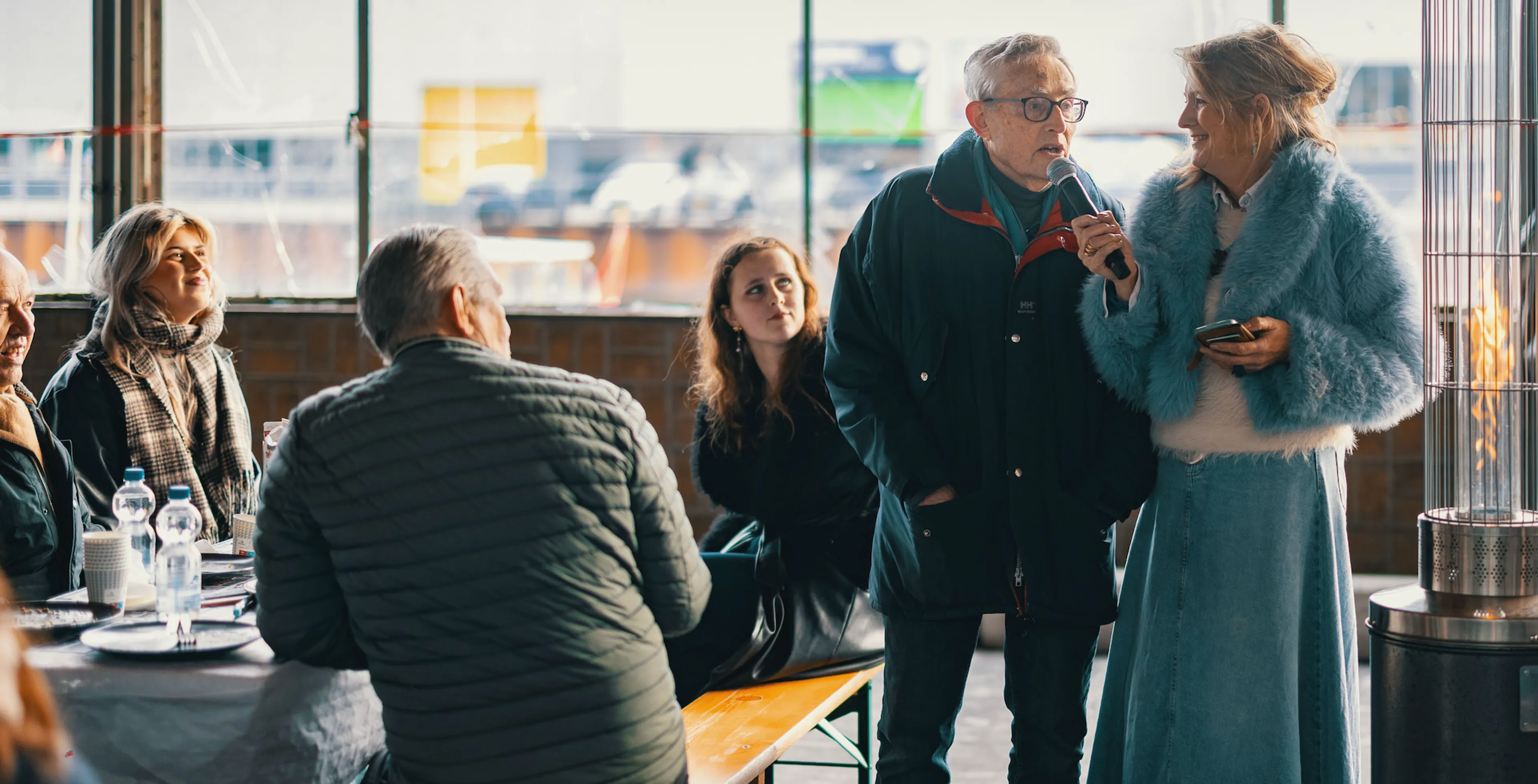 Twee mensen spreken voor een groep bezoekers in een lichte industriële ruimte, terwijl een terrasheater vlammen geeft.
