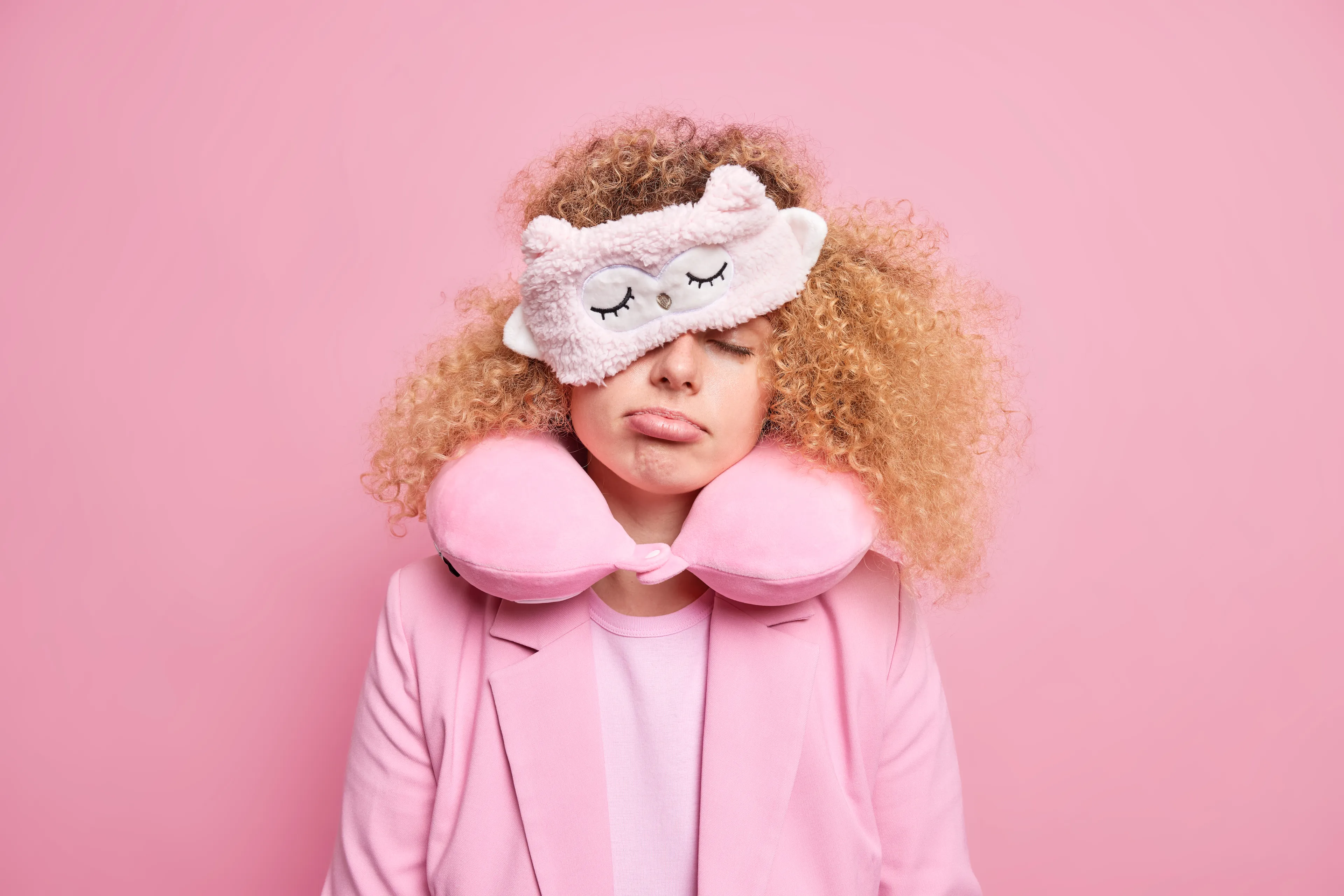 The portrait describes a young-appearing person with light blonde, curly hair, wearing a light pink blazer and shirt, a large U-shaped pink neck pillow, and a fluffy pink sleep mask with a stitched sleepy face and small ears slid over the eyes; the eyes are closed and the expression is tired and pouty, creating a sleepy, restful mood with a monochrome pink palette.