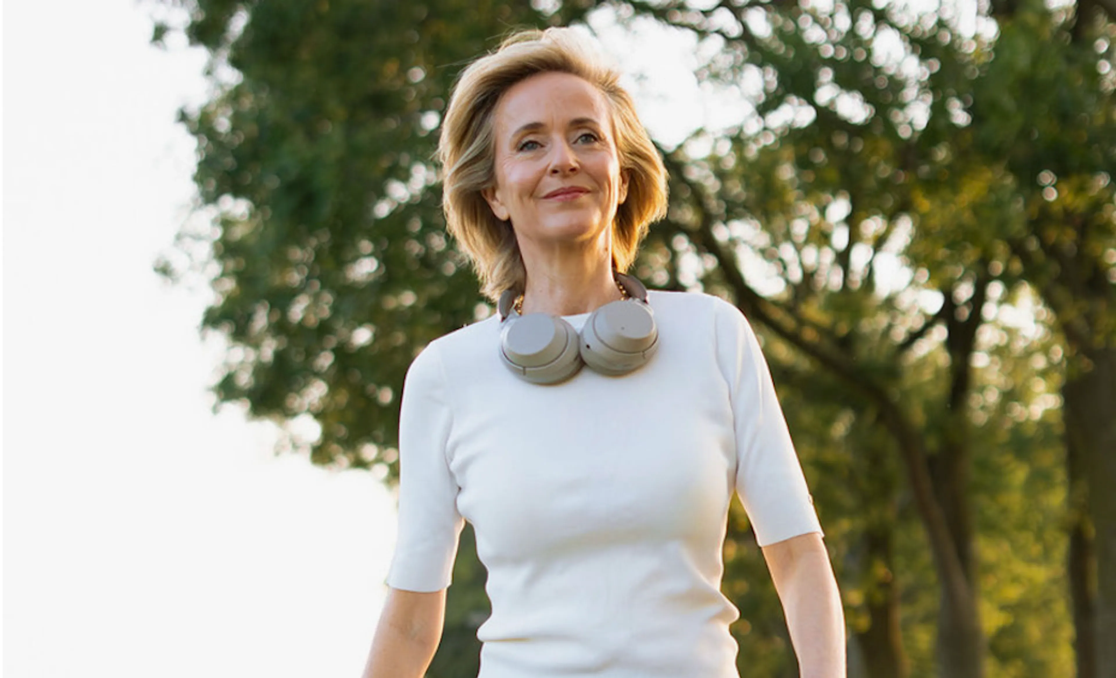 Onderwerp: een vrouw van middelbare leeftijd in een parkachtige omgeving. Ze loopt buiten, draagt een strak wit shirt en een grote grijze over-ear koptelefoon, en heeft een rustige glimlach terwijl ze omhoog kijkt; de achtergrond is een onscherpe, zonnige groenblauwe sfeer.
