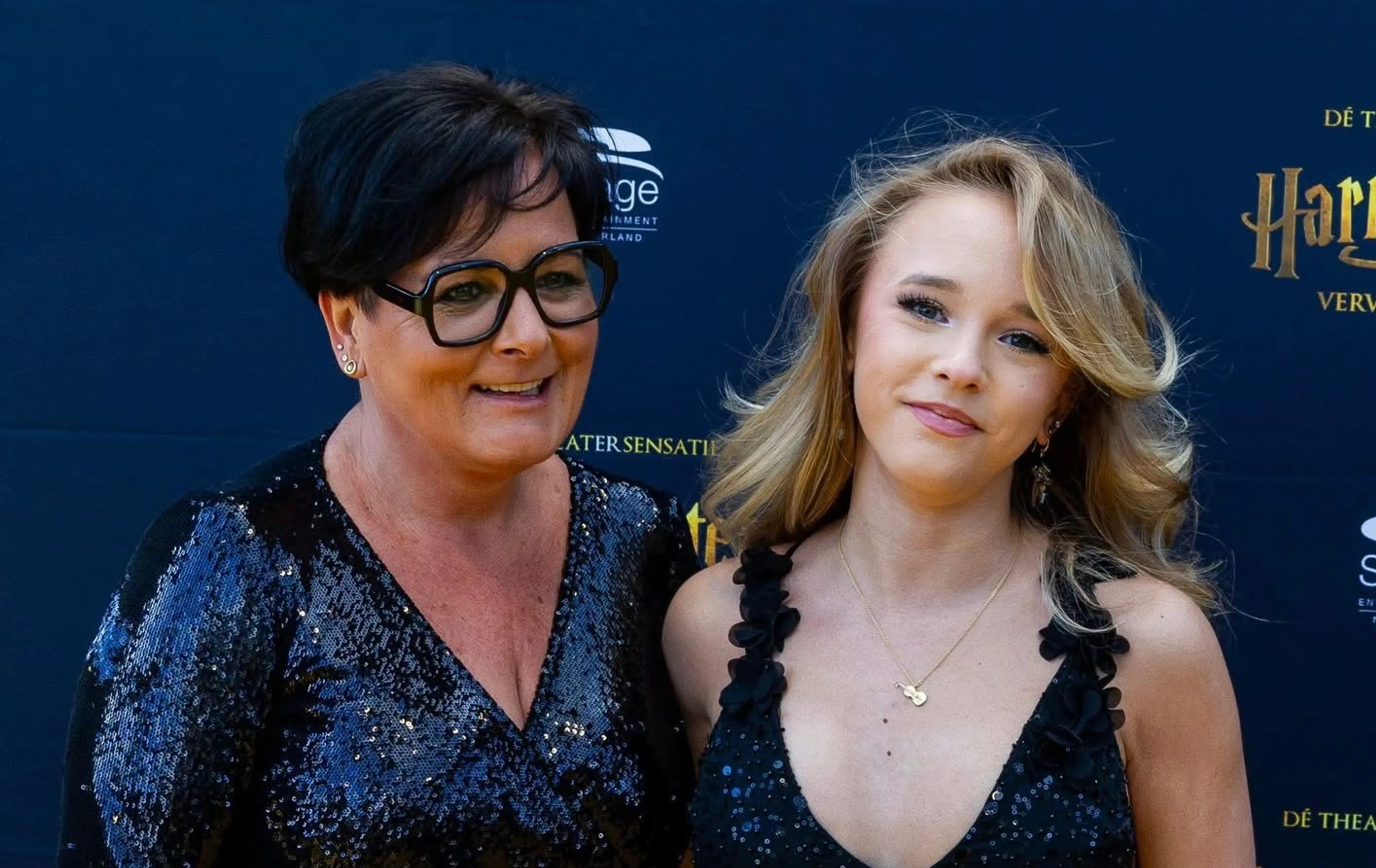 Harry Potter – De theatersensatie is de geafficheerde titel op een donkerblauwe step-and-repeat-achterwand vol logo’s en gouden bijschriften, waarmee een première- of persmoment wordt aangegeven. De foto toont twee vrouwen naast elkaar poseren: een oudere vrouw met kort donker haar en glinsterende top naast een jonge vrouw met lang blond haar en een sprankelend zwarte jurk.