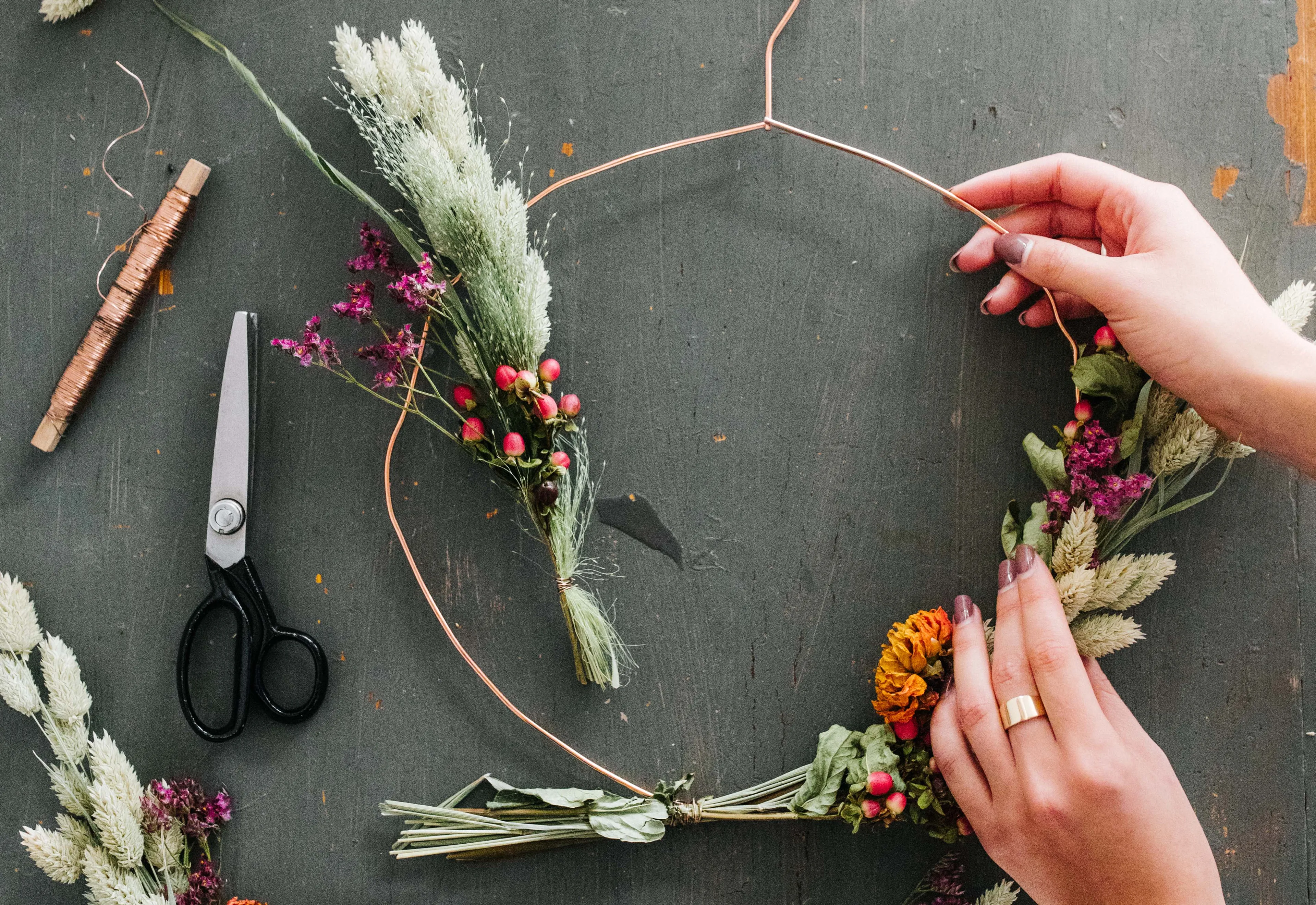 Op een werktafel zie je twee handen die om een boog ijzerdraad kersttakjes bevestigt. Op deze manier maakt ze een kerstversiering voor in huis.