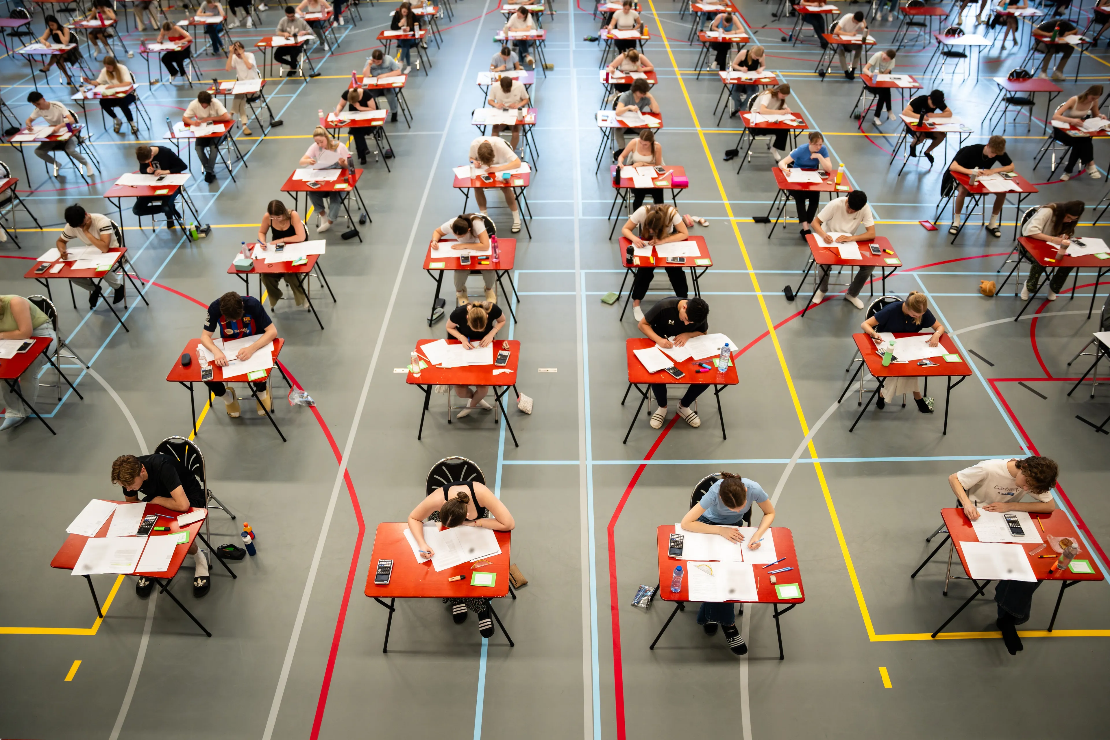 Scholieren maken hun examen in een gymzaal vol rode tafeltjes
