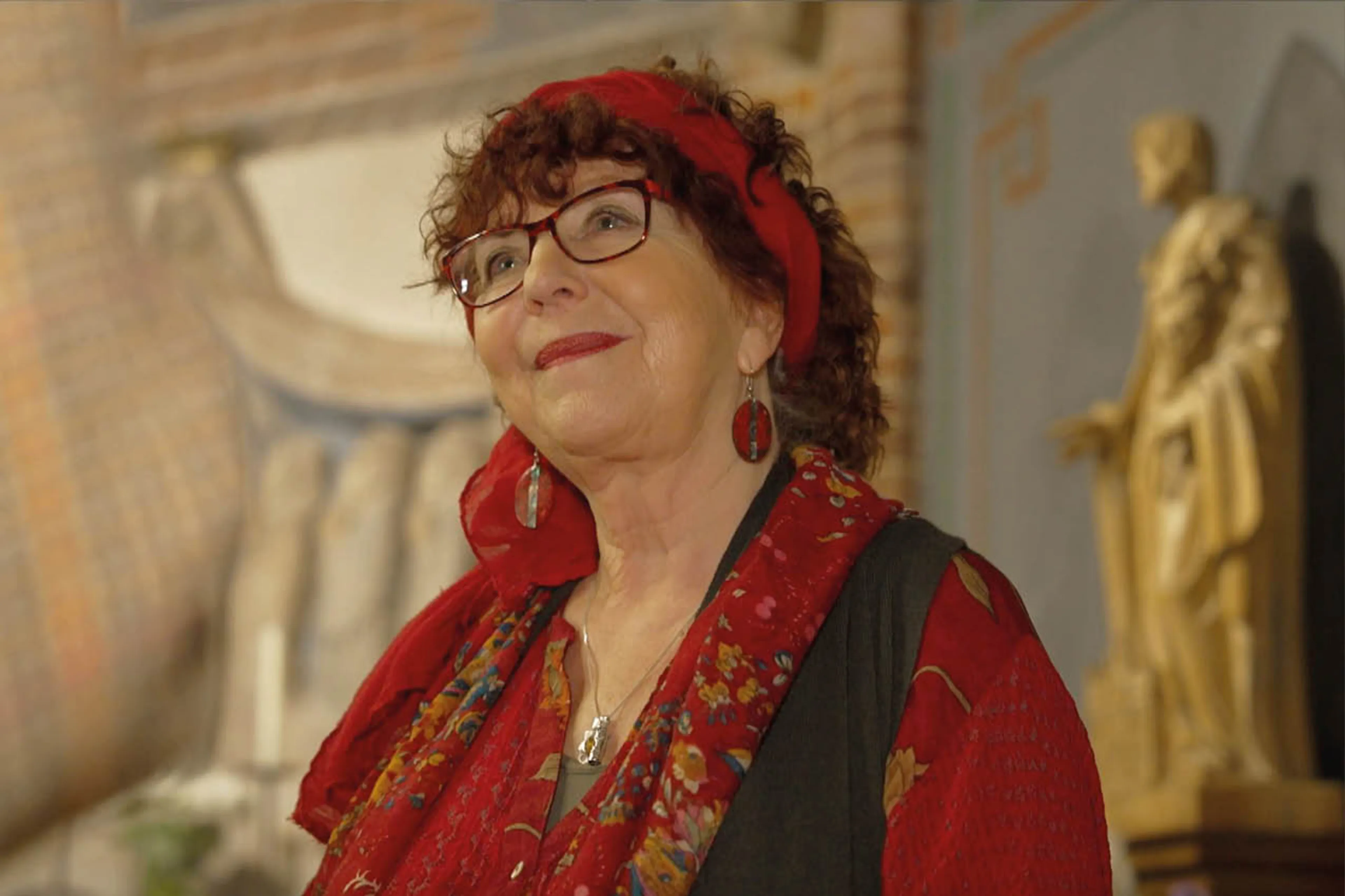 Elly Zuiderveld kijkt omhoog in een kerk. Ze heeft rode kleren aan, en draagt een sjaal in haar krullende rode haren. Haar lippen zijn rood gestift.