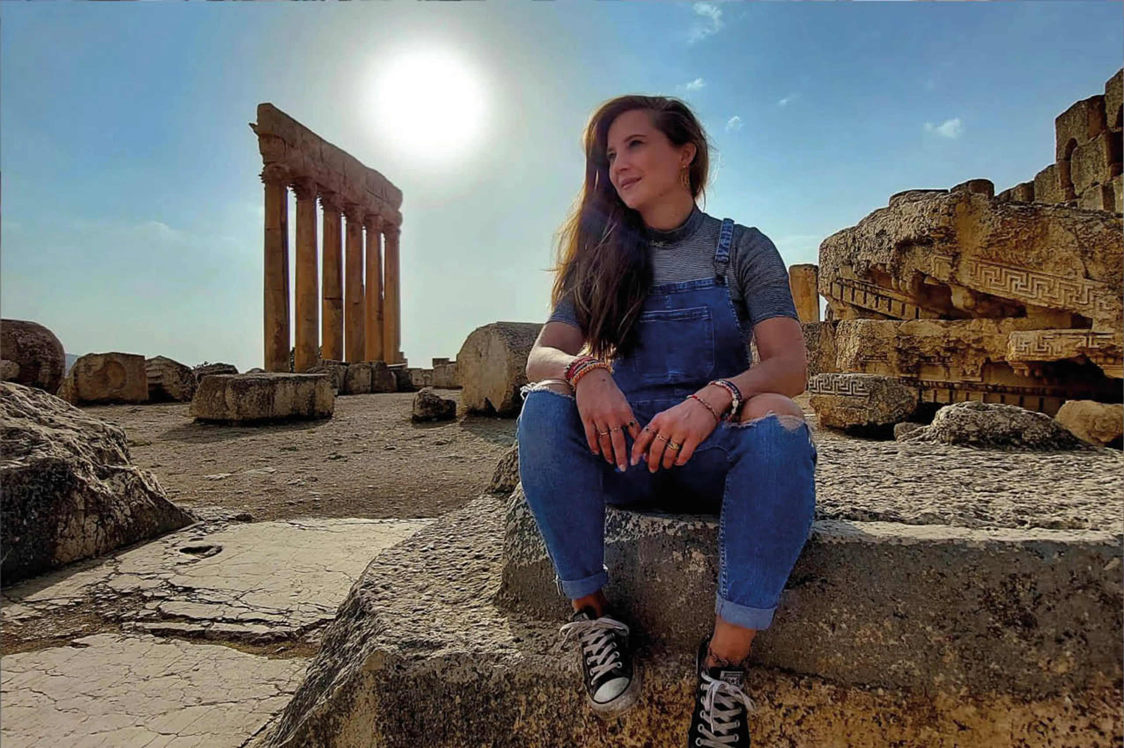 Mirjam zit in haar tuinbroek op een rots in de zon. Ze heeft lange bruine haren over een kant van haar schouder. Achter haar zie je een rotsachtige omgeving en een oude ruïne.