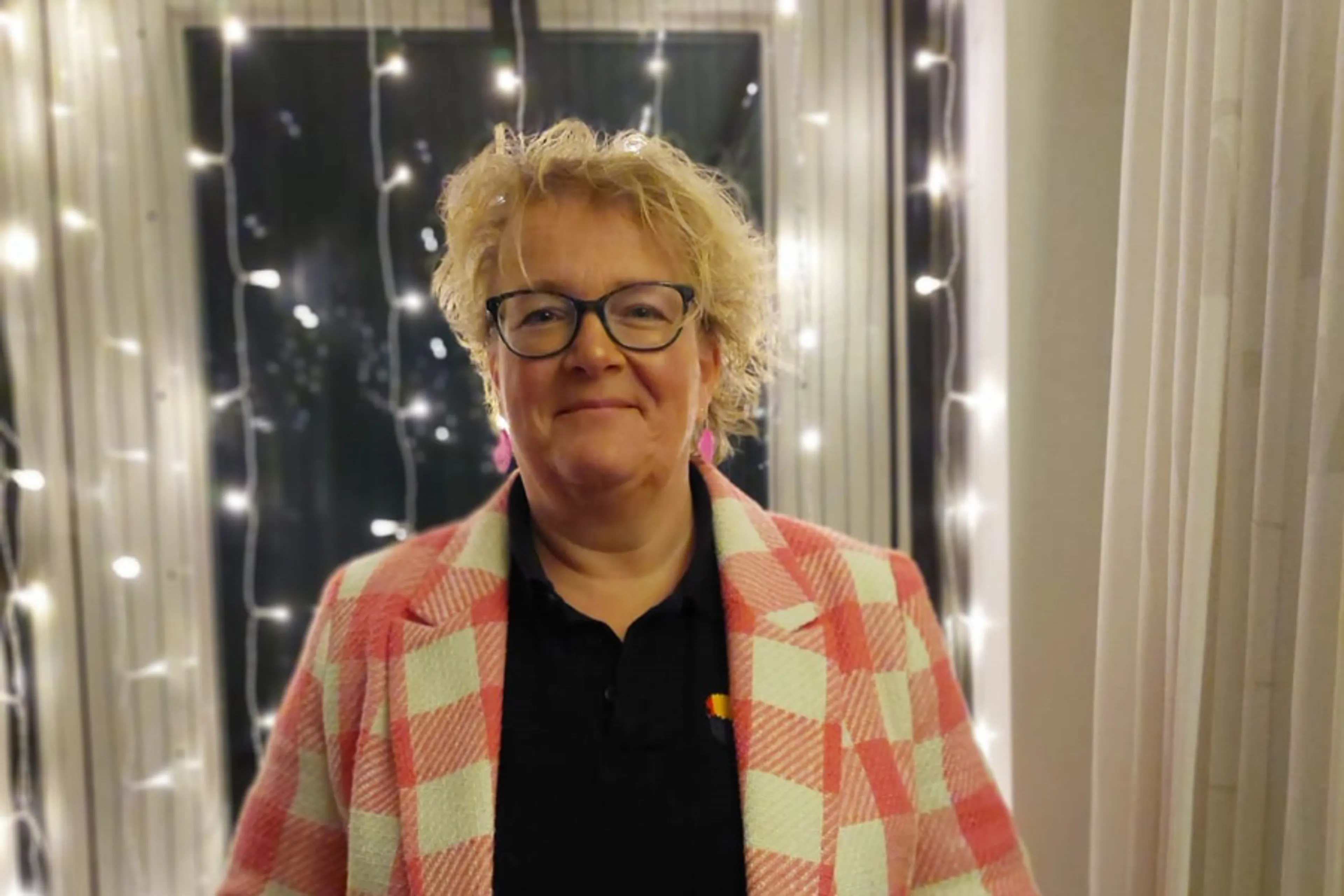 Willemien staat voor een raam met lichtjes. Ze heeft blond kort slag haar en een bril op. Ze glimlacht in de camera en draagt een rood-wit geblokt jasje met een zwart t-shirtje eronder.