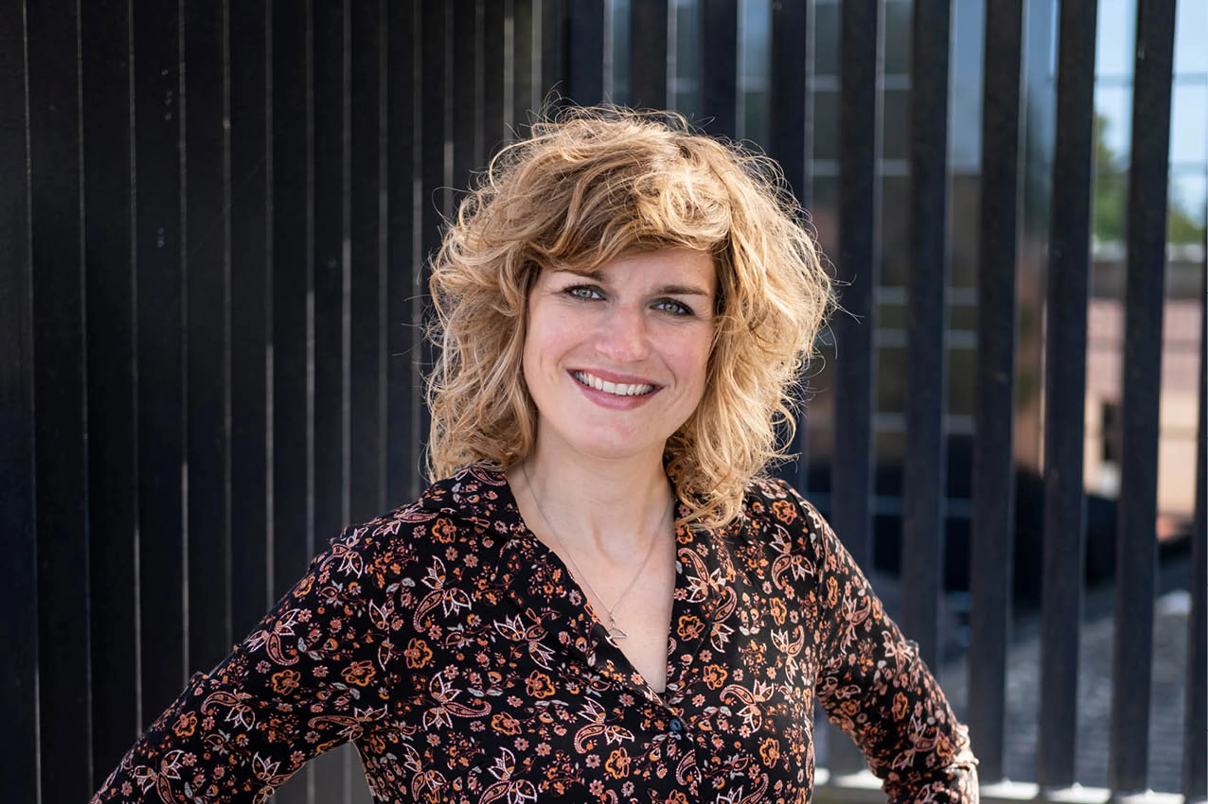 Stylecoach Maria staat voor een hek, ze draagt een bloemenjurk en lacht naar de camera.