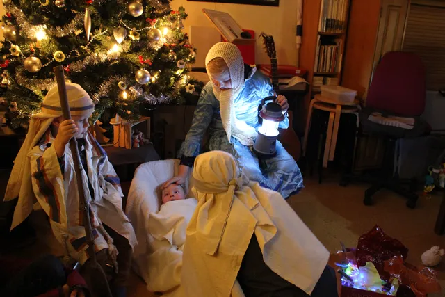 Bij een verlichte kerstboom spelen drie kinderen het kerstverhaal na. Er ligt een baby in een kribbe en er staan drie herders omheen.