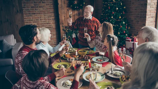 Tafel vol eten en drinken. Aan de tafel zitten kinderen, ouders en grootouders. Ze hebben een kersttrui aan en toasten met hun glas.