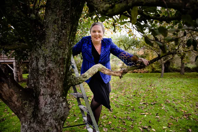 Dineke staat bij een boom en lacht in de camera. Ze heeft een paarsblauw jasje aan met een zwart jurkje. Haar lippen zijn rood gestift en haar haren zitten in een knotje.