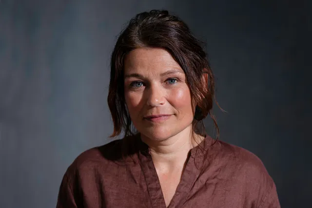 Marijke staat voor een donkere achtergrond en met studiolicht op haar gezicht. Ze heeft haar handen rond haar buik gesloten op elkaar liggen.