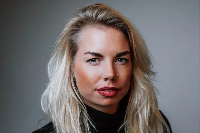 Op de foto staat Gloria, ze groeide op als KOPP/KOV-kind. Ze heeft blond haar, felrode lipstick en kijkt direct de camera in.