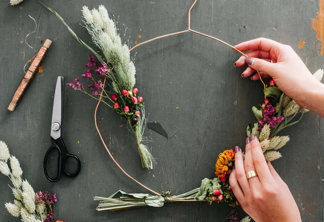 Op een werktafel zie je twee handen die om een boog ijzerdraad kersttakjes bevestigt. Op deze manier maakt ze een kerstversiering voor in huis.