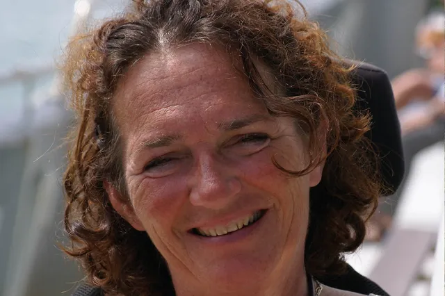 Rianne lacht met een grote glimlach in de camera. Ze heeft kort bruin krulletjes haar.