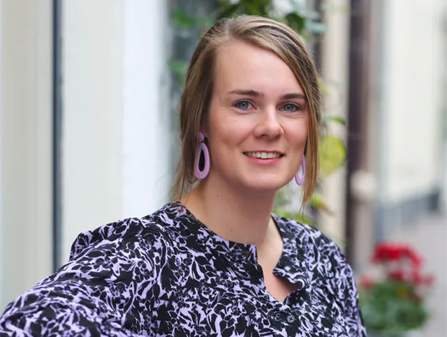 Op de afbeelding zit schrijfster Leanne Pots met een paarse jurk met zwarte print voor een witte muur op een bankje. Ze kijkt lachend naar de camera en heeft haar bruine haar half opgestoken.