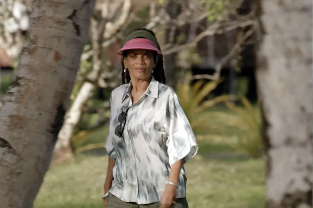 Presentatrice Aldith Hunkar staat met een petje en een blouse tussen twee bomen voor het EO programma 'Firma Erfgoed Overzee'.