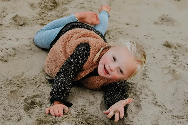 Livia ligt in het zand. Ze lacht naar de camera en steekt haar hand uit.