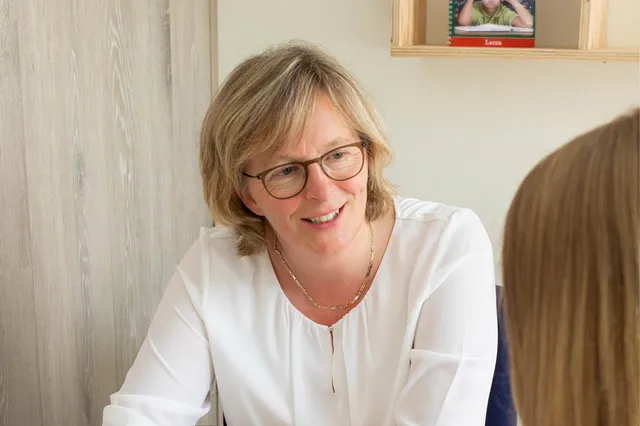 Sandra heeft een wit shirt aan, kort donkerblond haar en een bril. Ze lacht naar haar client die tegenover haar zit.