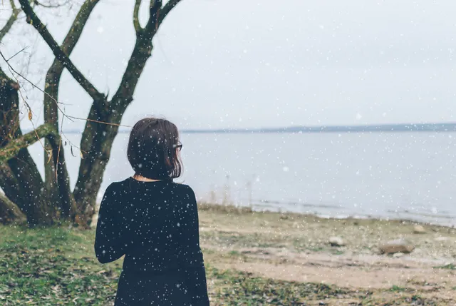 Vrouw met kort bruin haar, kijkt over het water. Ze heeft een zwarte jurk aan. Ze kijkt uit op wat zand, wijd water en naast haar staat een kale boom.