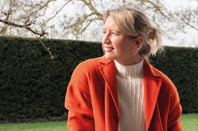 Hanneke Bouman in de tuin, met haar blonde haren in een knotje. Ze kijkt weg van de camera, maar glimlacht wel. Haar oranje stoffen jas heeft ze open en daaronder draagt ze een gebreide coltrui van gebroken wit.