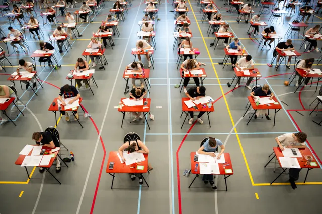 Scholieren maken hun examen in een gymzaal vol rode tafeltjes.