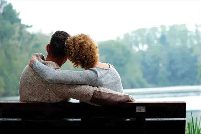 Koppel op bankje, armen om elkaar heen. Vrouw heet een grijsblauwe trui aan met oranje krulletjeshaar. Man heeft grijze trui met kort donker haar. Ze kijken naar het water voor zich.