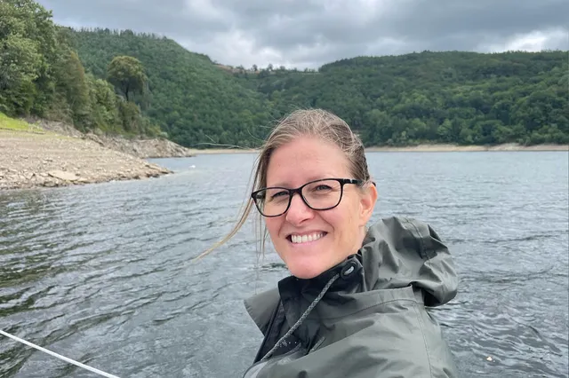 Marian staat in het buitenland bij het water. Ze draagt een kakigroene regenjas met capuchon, een zwarte bril en haar haren in een staart. Ze lacht in de camera met knijpende oogjes tegen het licht. Achter haar zie je donkergrijze wolken en groene bergen.