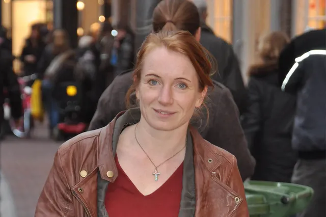 Bernadette van Dijk heeft rossig haar in een paardenstaart gebonden. Ze draagt een bruin leren jack en een ketting met een kruisje om haar hals.