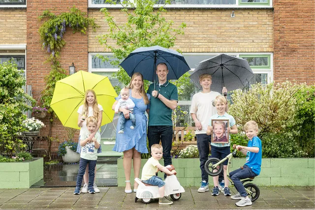 De familie Blom staat onder drie paraplu's voor hun huis. Er staan naast de ouders zeven kinderen. Een van hun zoons heeft een foto vast van hun overleden dochter Mirjam.