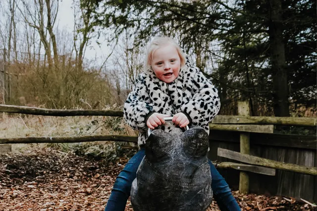 Livia zit met haar benen aan weerszijden op een speeltoestel. Ze draagt een wit met zwart gevlekt jasje en lacht in de camera. Op haar wangen is een rode blos te zien.