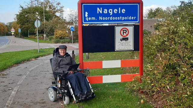 Op de foto zit Ab in zijn rolstoel voor een verkeersbord dat het begin aangeeft van het dorp Nagele.