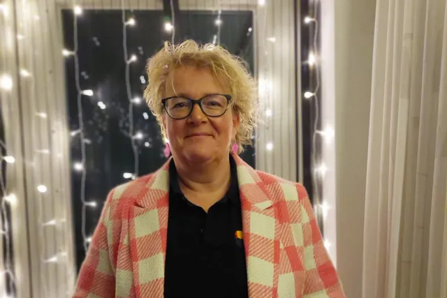 Willemien staat voor een raam met lichtjes. Ze heeft blond kort slag haar en een bril op. Ze glimlacht in de camera en draagt een rood-wit geblokt jasje met een zwart t-shirtje eronder.