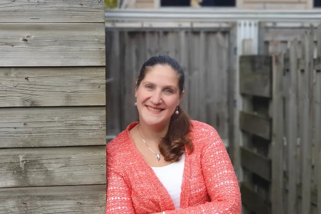 Silvia leunt tegen een schuurtje aan, lacht in de camera en heeft een roze vestje aan.