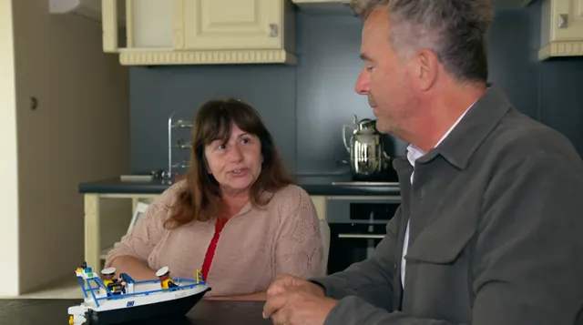 Foto van Engel uit 'Het Familiediner' met Bert van Leeuwen. Engel is een vrouw van middelbare leeftijd, heeft bruin haar met een pony. Ze draagt een beige blouse met een rood hemdje eronder. Ze zit in de keuken met Bert. Voor haar op tafel staat een speelgoedboot.