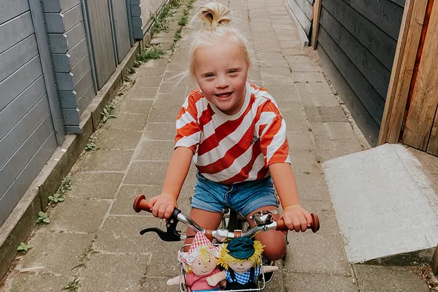 Livia zit op de fiets, ze lacht naar de camera. Voor op haar fiets in een mandje zitten twee knuffels.
