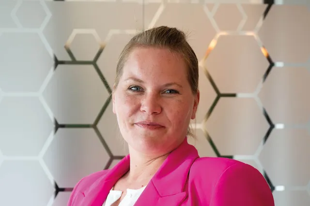 Wendy kijkt in de camera en glimlacht. Ze draagt een felroze blazer en haar haren strak naar achteren in een staart.