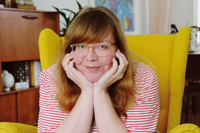 Marloes is kunstenaar. Ze draagt een rood-wit gestreept shirt, heeft een bril op haar hoofd, heeft rossig halflang haar met een pony en leunt met haar kin op haar handen. Je ziet een deel van de okergele stoel en haar jaren 60 kast.