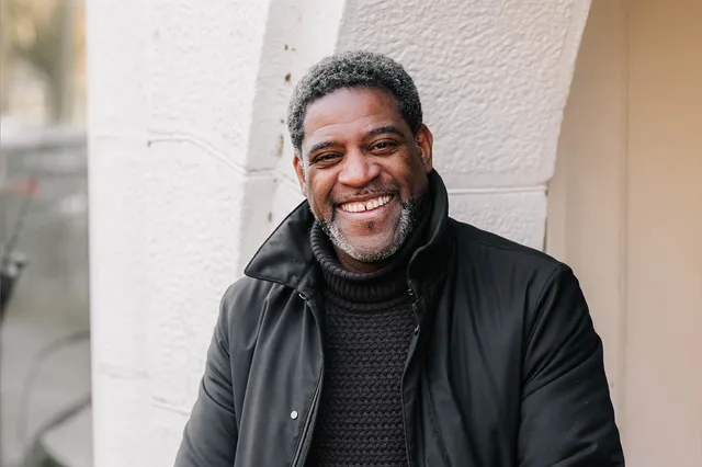 Televisiepersoonlijkheid Sergio Vyent heeft een zwarte jas aan met een donkergrijze coltrui eronder. Hij lacht in de camera, waardoor er een klein spleetje tussen zijn witte tanden zichtbaar wordt. Zijn zwarte kroeshaar is langzaam aan het vergrijzen.