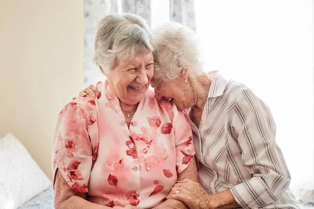 Foto van twee oude vrouwen die lachend bij elkaar zitten.