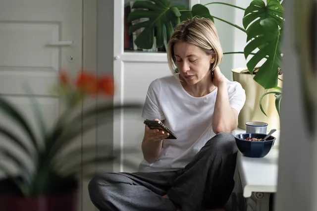 Vrouw van middelbare leeftijd kijkt op haar telefoon. Ze heeft een wit shirt aan en een grijze broek. Haar korte blonde haren zitten achter haar oor. Naast haar staat een kommetje met eten.