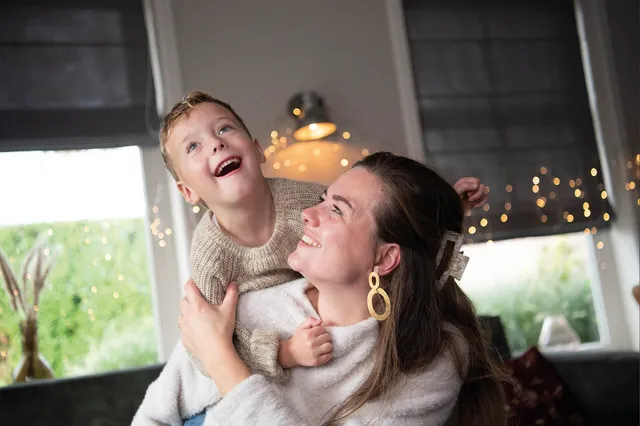 Moeder Anne Christine met haar zoon Sion. Ze lachen naar elkaar en er zijn gezellige lichtjes op de achtergrond.