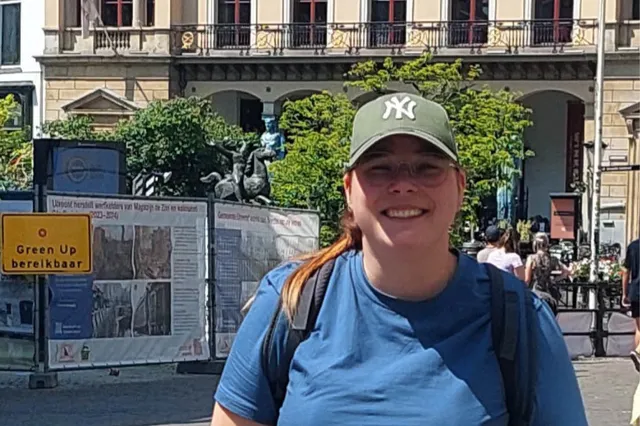 Sarina staat in de stad met een petje op, lacht naar de camera. Haar koperkleurige haar zit in een paardenstaart, en ze heeft een rugzak op haar rug.