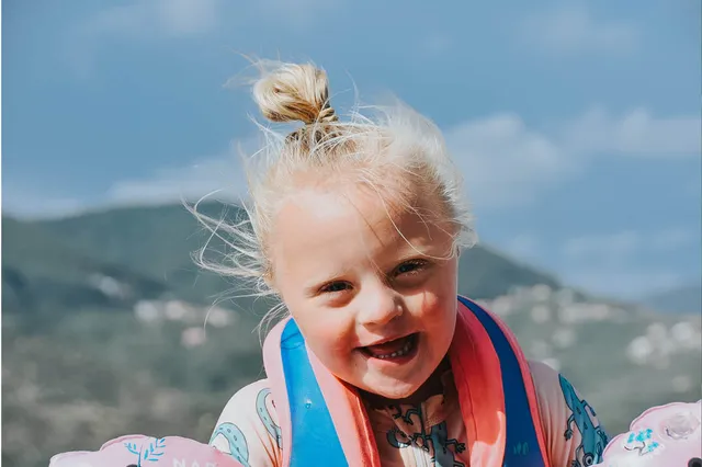 Livia heeft haar witblonde haren een knotje en een zwemvest aan. Achter haar zie je de Italiaanse bergen. Ze lacht breeduit in de camera.