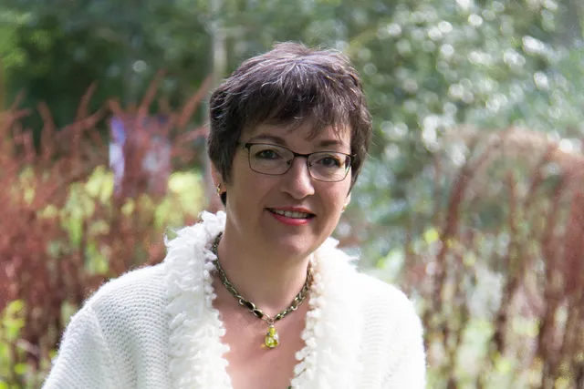 Op de foto staat Marian in een tuin. Ze draagt een bril en heeft kort haar. Ze draagt een wit vest met een opvallende witte kraag en een ketting.