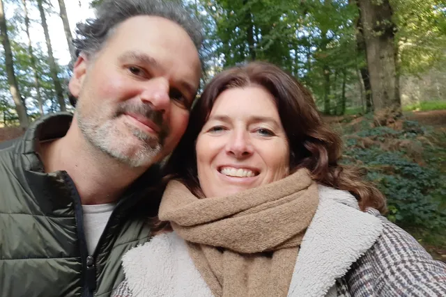 Marlies en Marc lachen breeduit in de camera. Het is herfst, ze hebben beiden een jas aan en staan dicht bij elkaar.