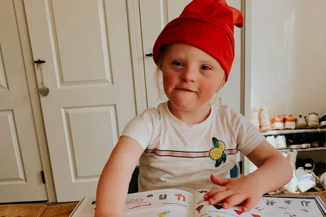 Livia, de dochter van Mirjam, heeft het Syndroom van Down en leest een boek met een kaboutermuts op terwijl ze speels de camera in kijkt.