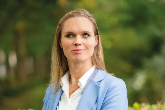 Tjarda Struik heeft blonde halflange haren met blauw-groene ogen en glimlacht trots in de camera. Over haar witte blouse draagt ze een blauw net jasje. Ze staat buiten in de groene natuur.