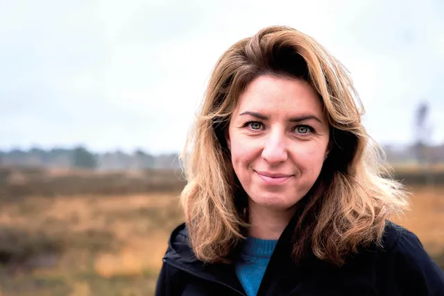 Joanna glimlacht in de camera. Ze heeft blond haar tot op haar schouders en blauwe ogen. Op de achtergrond is de heide te zien.