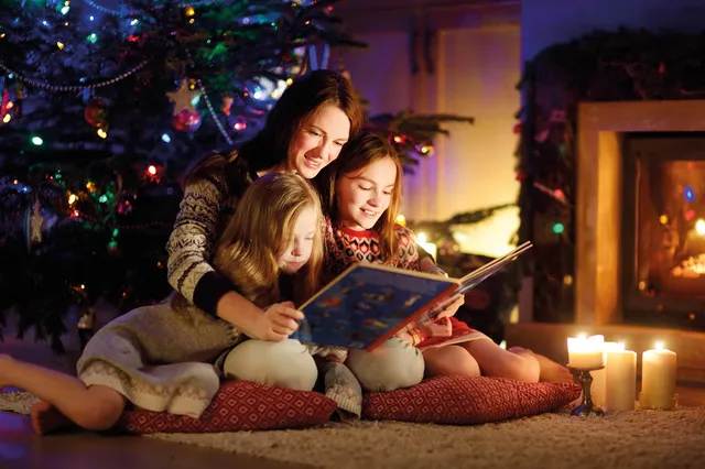 Vrouw met twee kinderen leest een verhaal voor bij de kerstboom en het haardvuur.