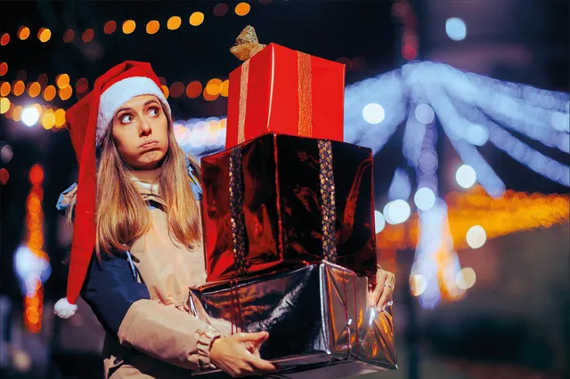 Stockfoto van jonge vrouw met blond haar en een kerstmuts op. Ze staat buiten bij een kerstmarkt met een stapel cadeaus in haar handen en kijkt zwoegend.